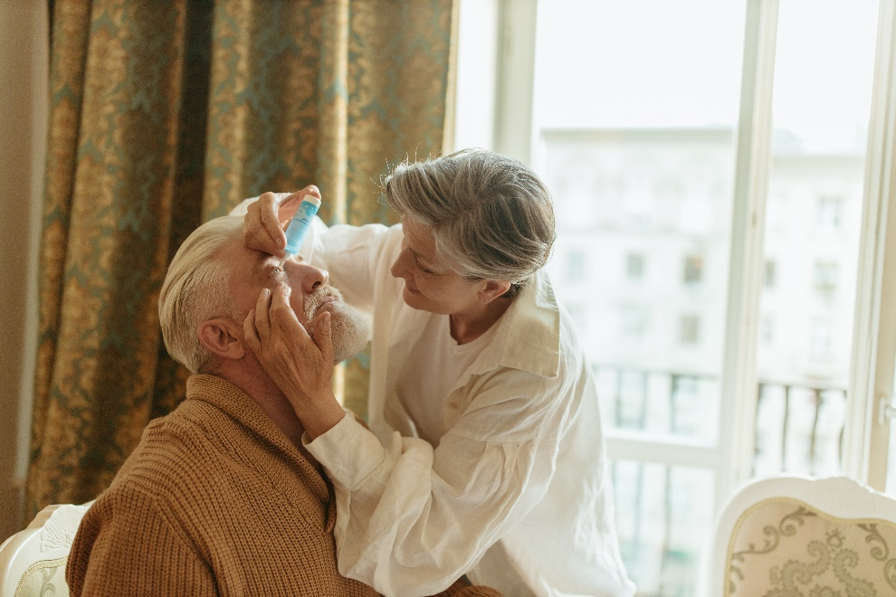 An elderly woman putting eye drops using a dropper in the eyes of an elderly man