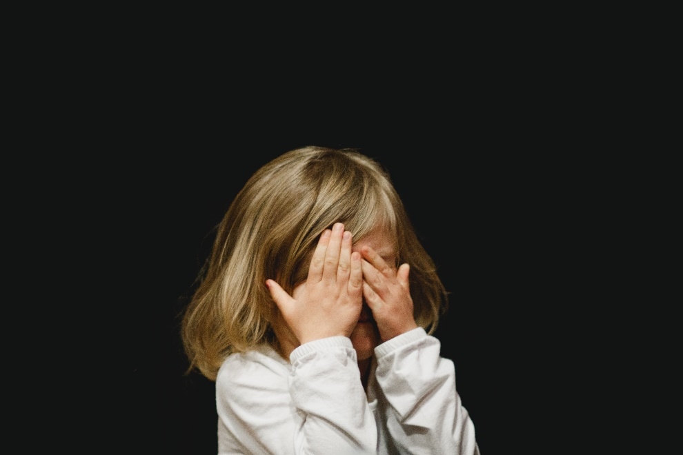 A little girl wearing a white shirt covering her eyes with her hands