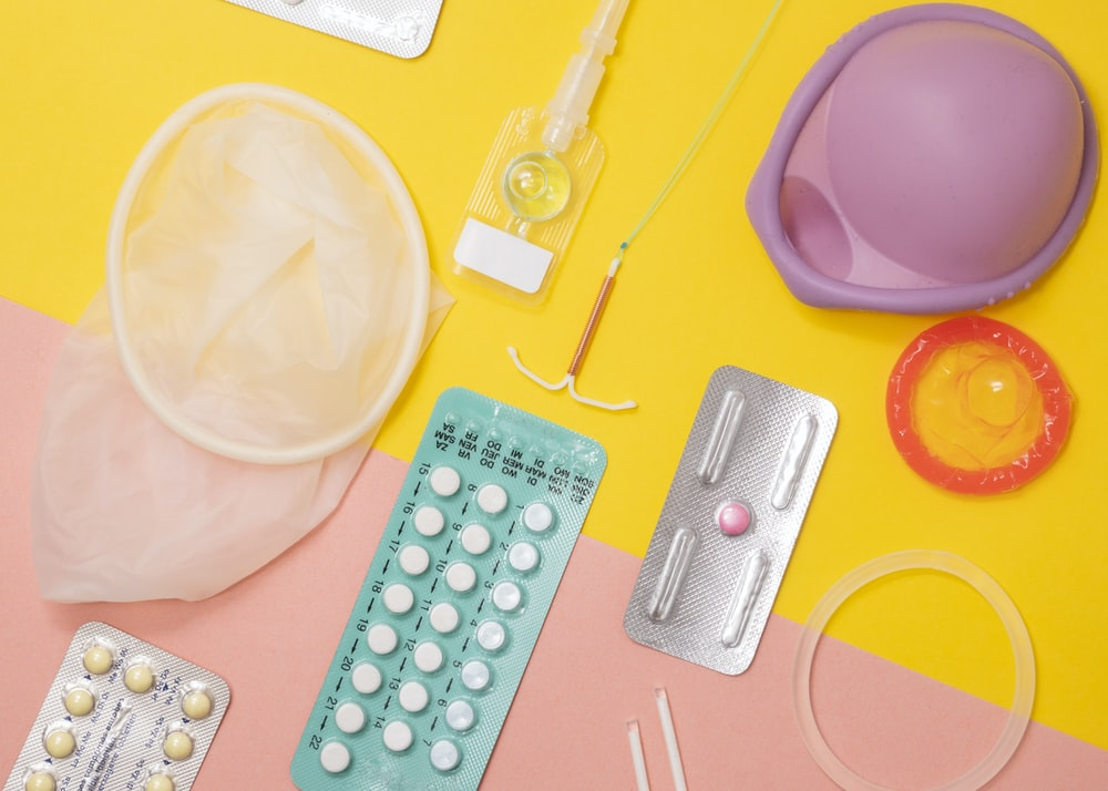 A range of contraceptive methods lying on a surface, including contraceptive pills, emergency contraception, condom, IUD, vaginal ring, implant