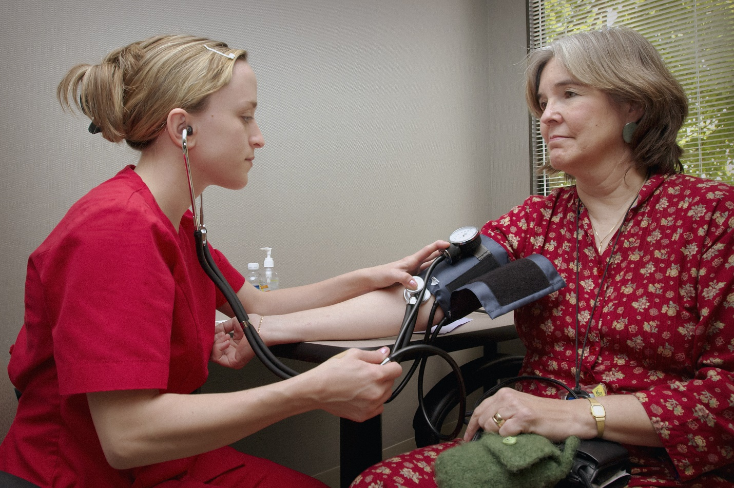 A doctor wearing red scrubs using a stethoscope to check her patient's blood pressure