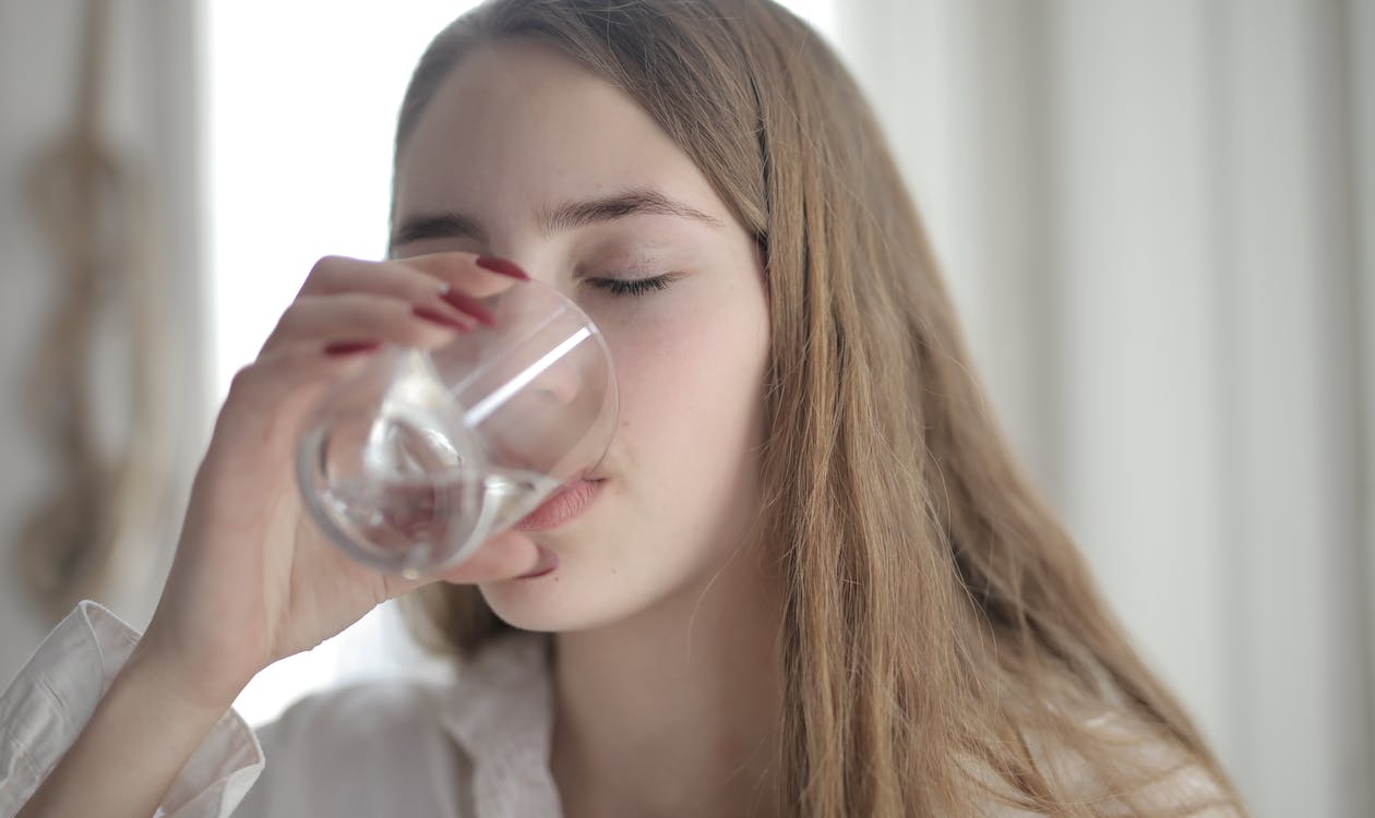A woman in a white shirt drinking water from a clear glass with her eyes closed