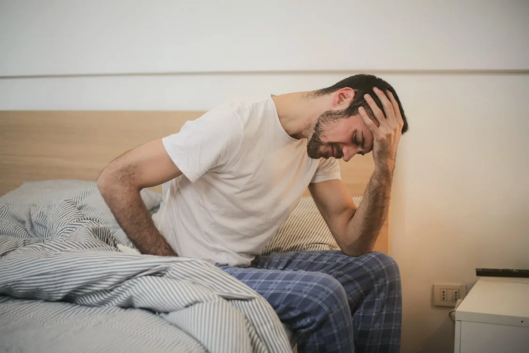 A photo of a man sitting on the bed and holding his head 