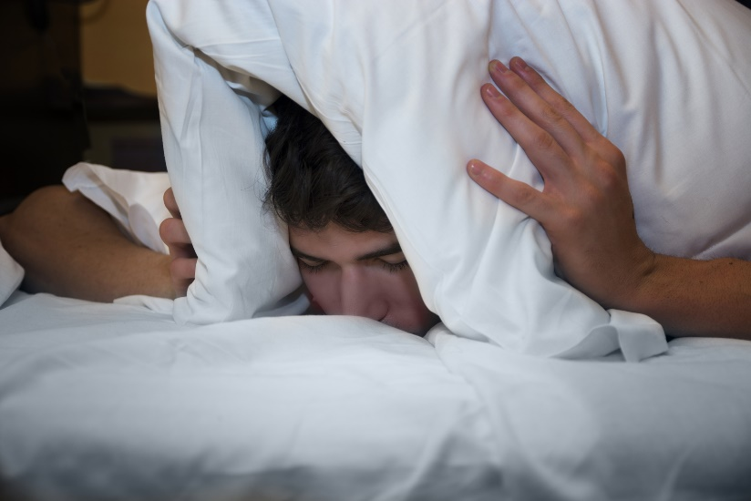 Sleep deprived man covering head with pillow.