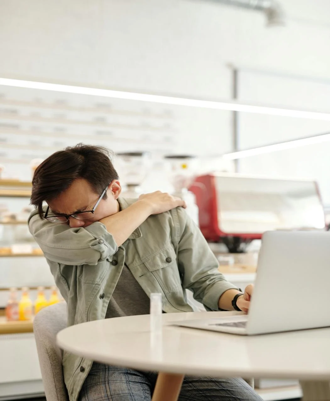 A man covering his mouth by his arm, while coughing, sitting in front of a laptop