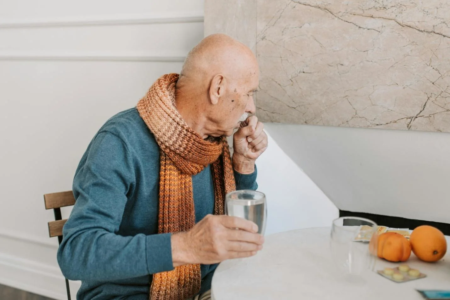 Elderly man coughing with a glass of water in his right hand while sitting on a chair