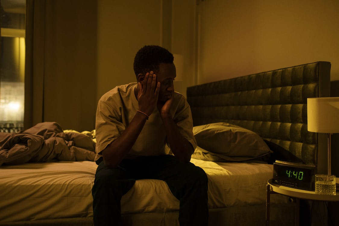 A man sitting on a bed, appearing uncomfortable due to urinary tract infection symptoms, considering an online doctor consultation.