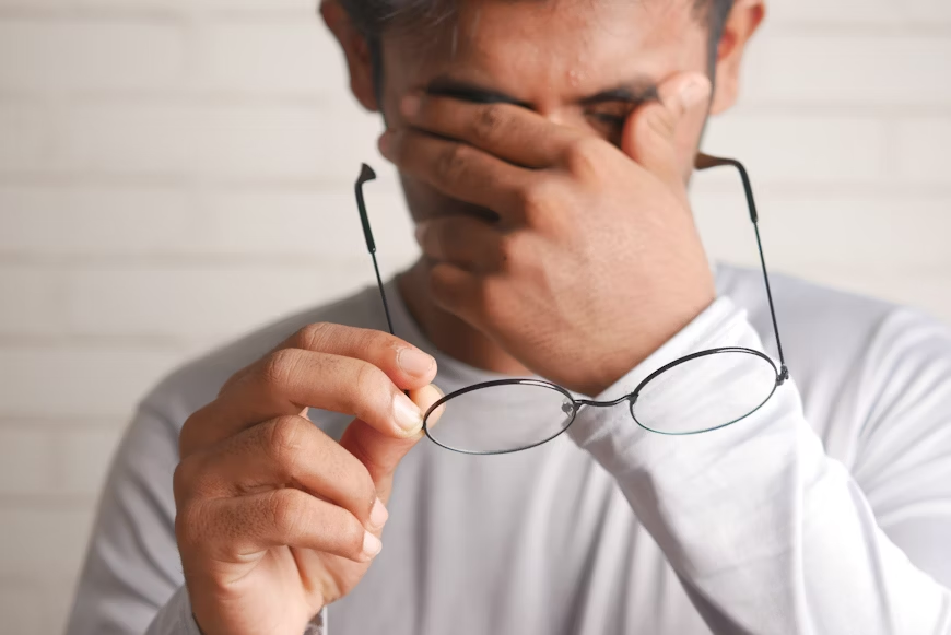 A man holding his glasses near his face, showing signs of eye irritation or pink eye discomfort.