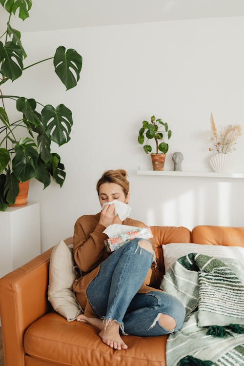 Woman sitting on a sofa and blowing her nose due to winter allergies.