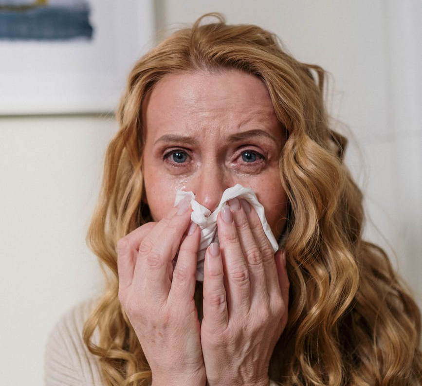 Woman blowing her nose, looking fatigued and uncomfortable.