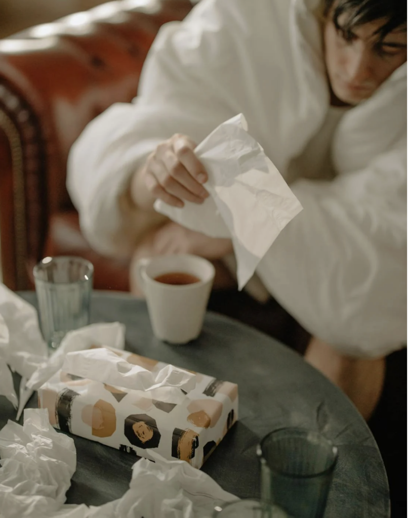 This photo shows a sick man sitting on a sofa beside tissues and tea.