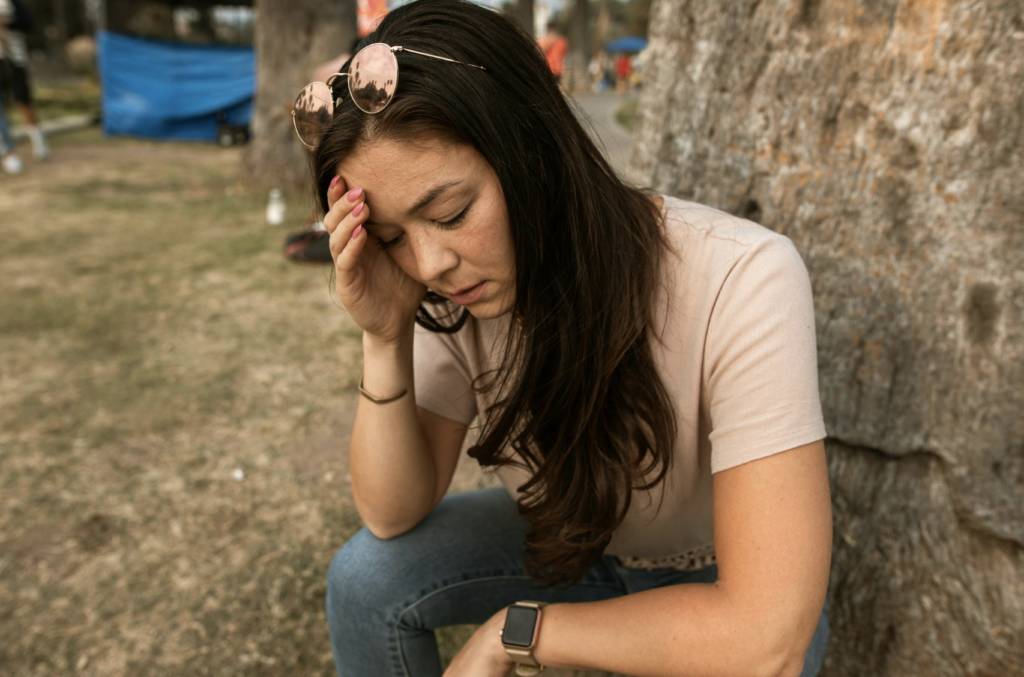 This photo shows a woman touching her forehead with her eyes closed and a tired expression outdoors.